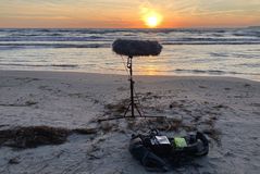 Im Vordergrund sieht man einen Mikrofon-Aufbau in einem Blimp als Windschutz auf einem Stativ. Im Hintergrund ist die Ostsee und der beginnende Aufgang der Sonne zu sehen.